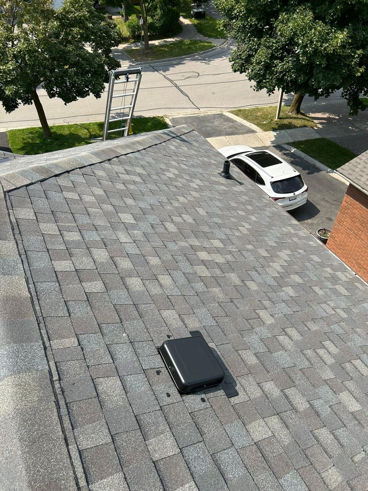View from a roof of asphalt shingles. A black vent and ladder are visible; a white car sits on the street.