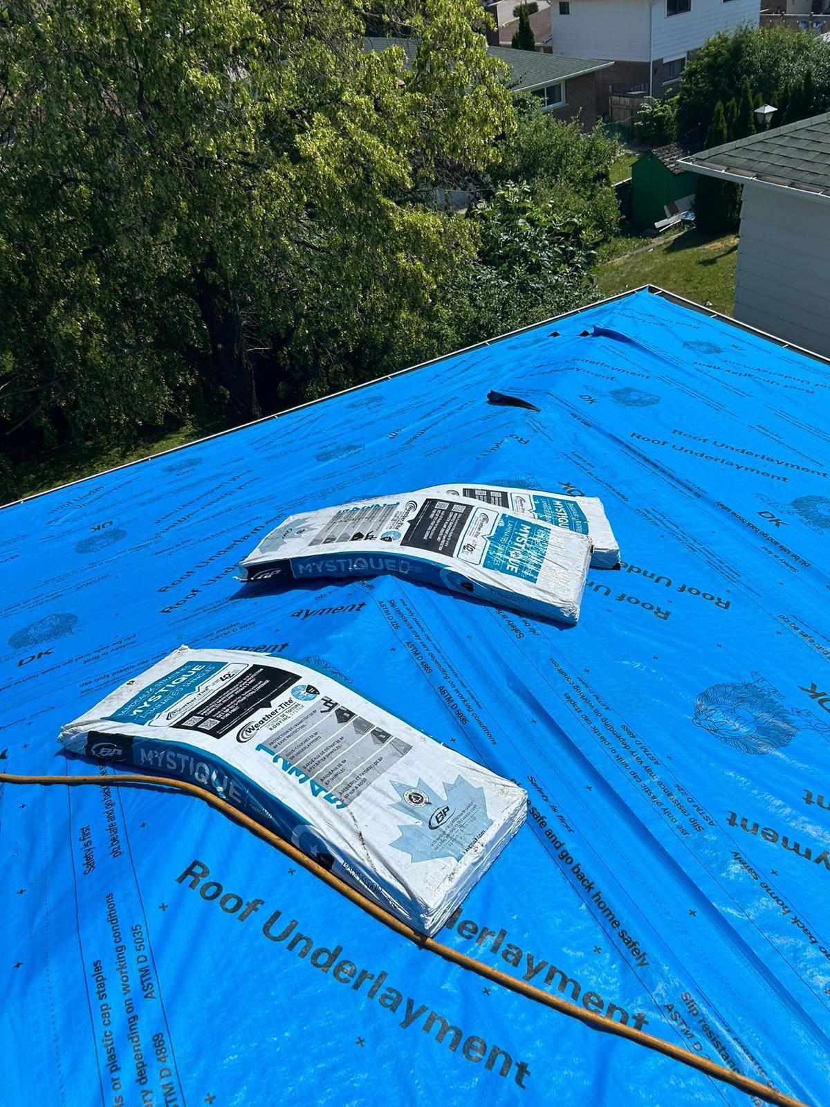 Blue tarp on a rooftop with multiple bundles of roofing shingles. Bright, sunny day, with trees in background.