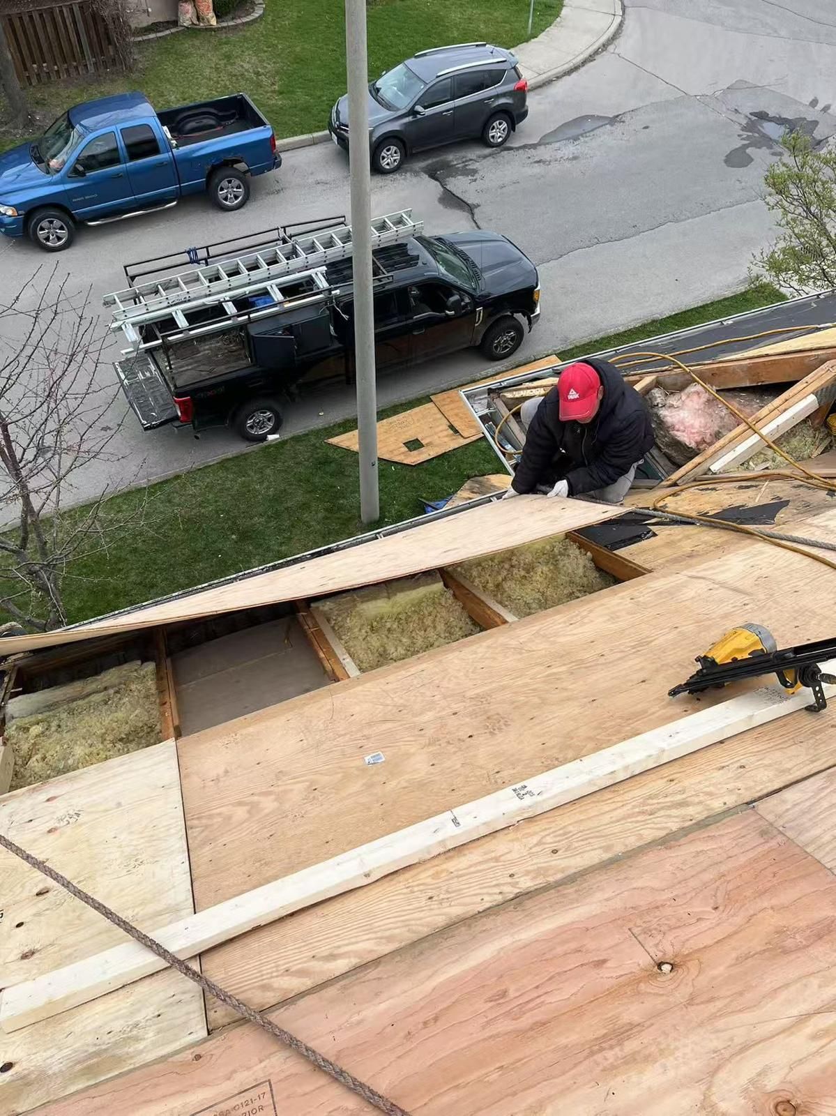 Roofer with red cap on roof installing plywood, surrounded by building materials. Trucks and street visible.
