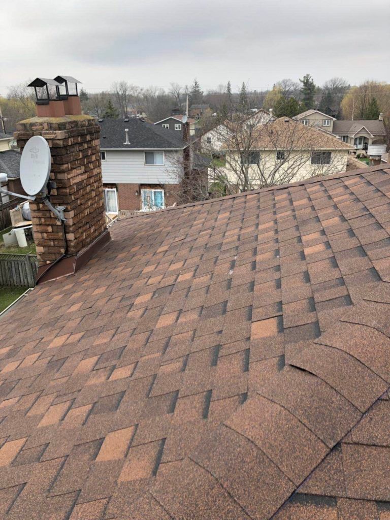 Brown shingled roof with a brick chimney and satellite dish; houses in background under overcast sky.