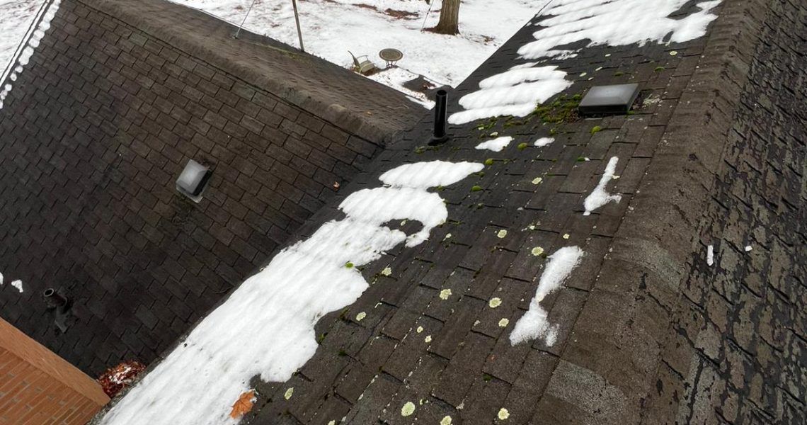 Dark shingled roof with patches of snow and a few vents, in a snowy setting.