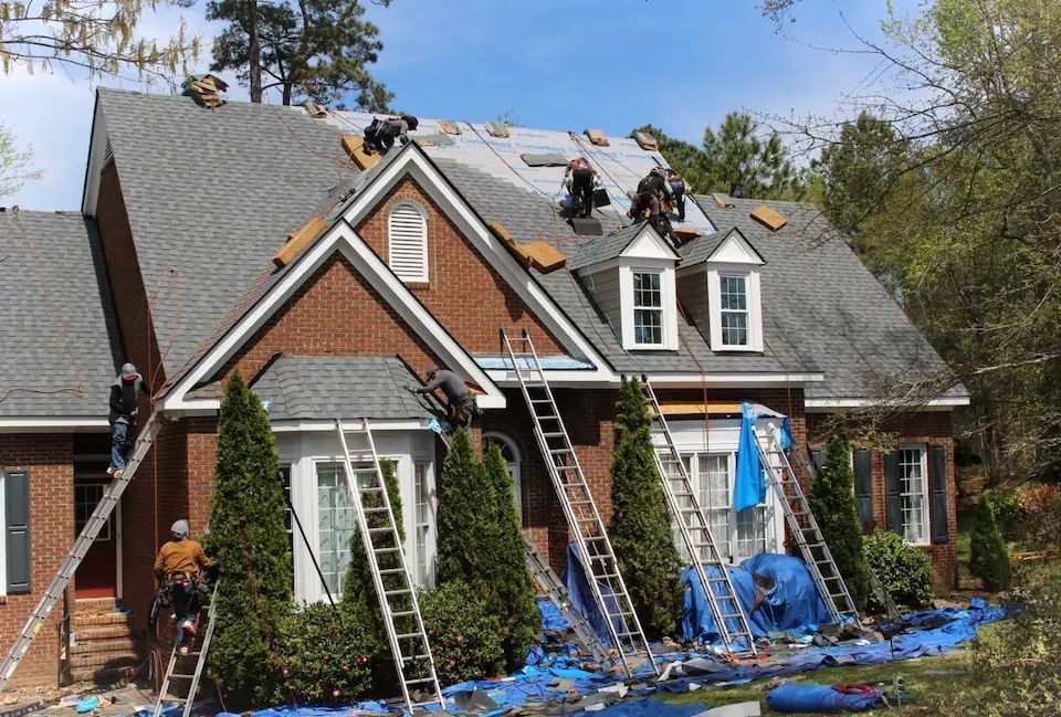 Roofers working on a brick house roof, using ladders, blue tarps, and shingles.