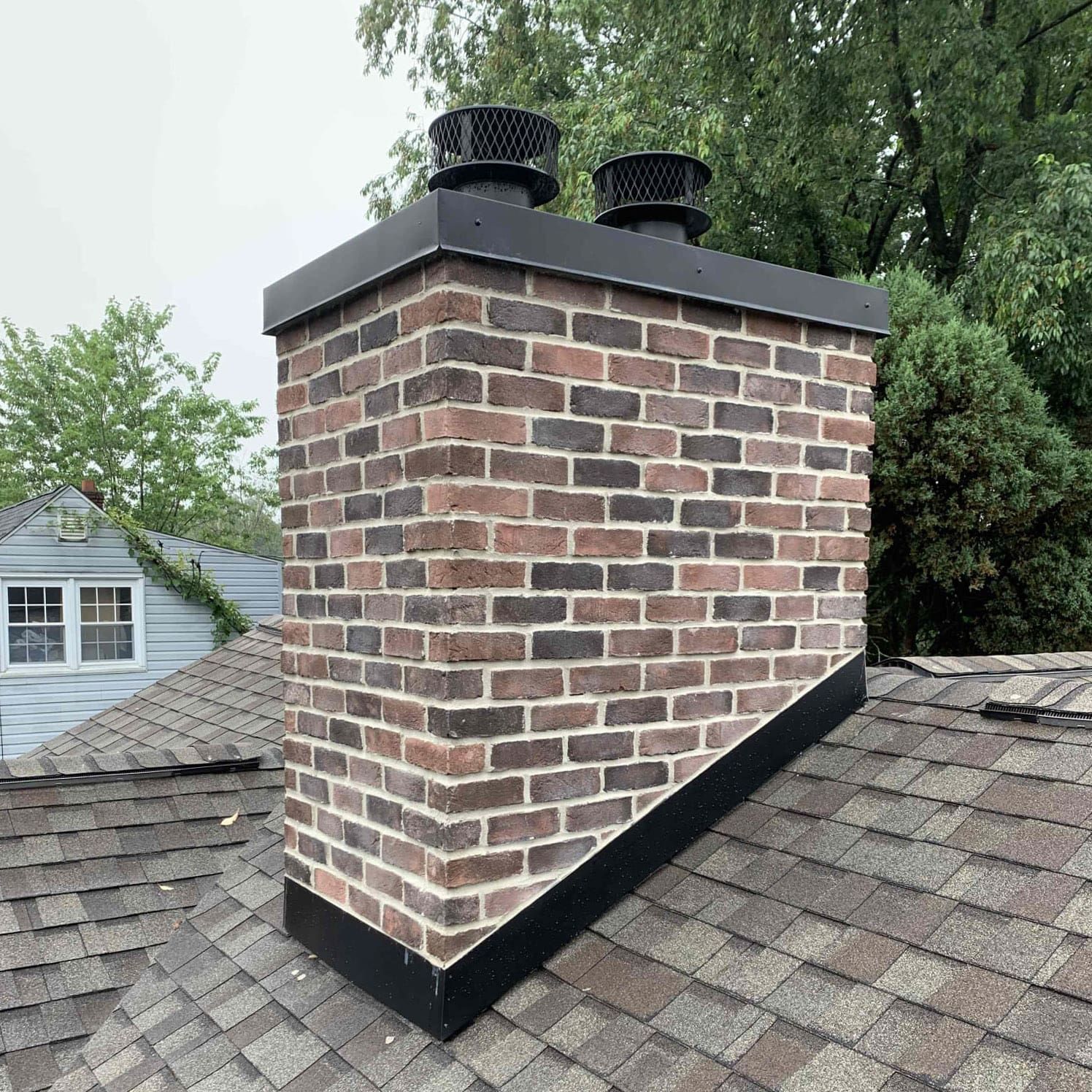 Brick chimney on a shingle roof with black metal trim and two chimney caps.