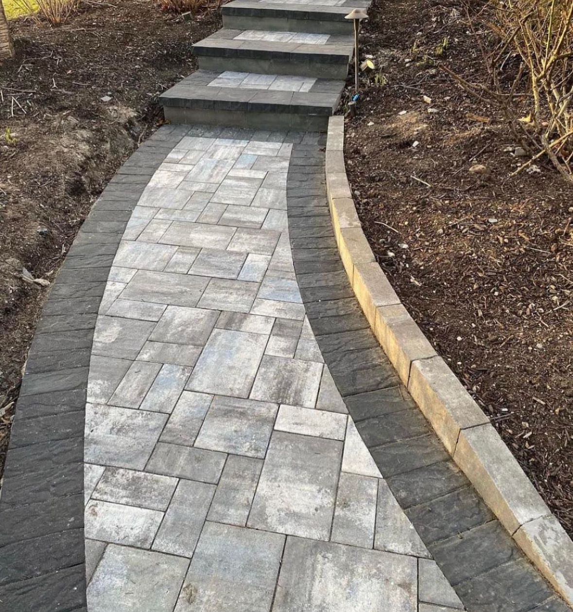 Brick pathway with dark border and steps leading uphill, alongside a dirt bed.