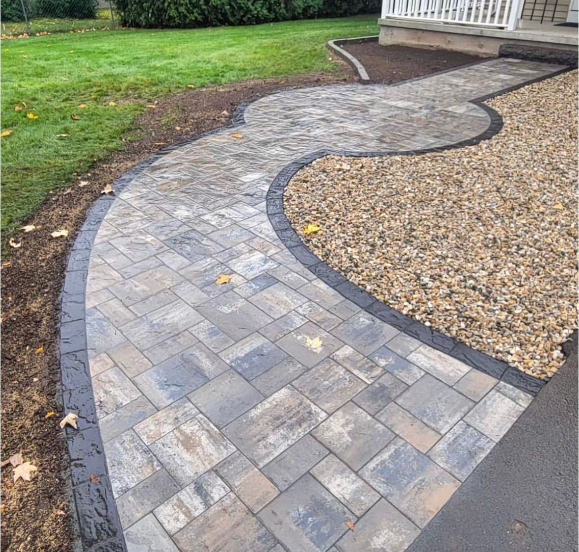 Brick walkway curves toward a house, bordered by dark edging and gravel. Green grass and driveway visible.