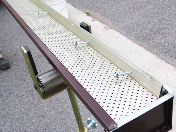 Brown gutter with perforated cover secured with bolts on a metal stand outdoors.