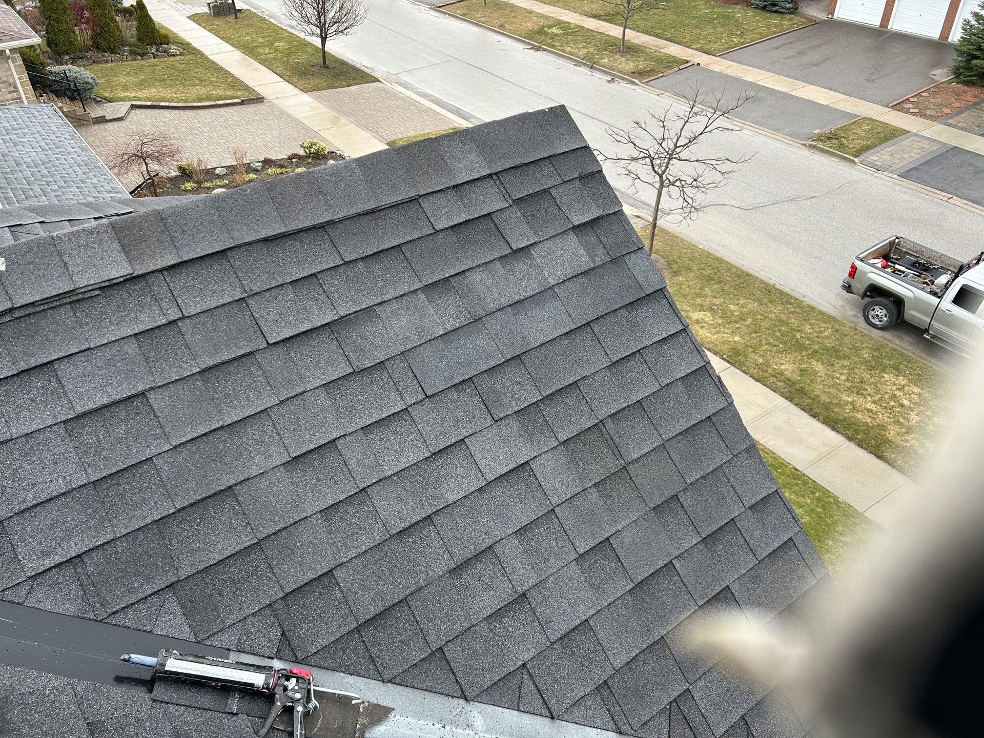 Gray asphalt shingles on a roof, viewed from above, with a street and parked truck in the background.