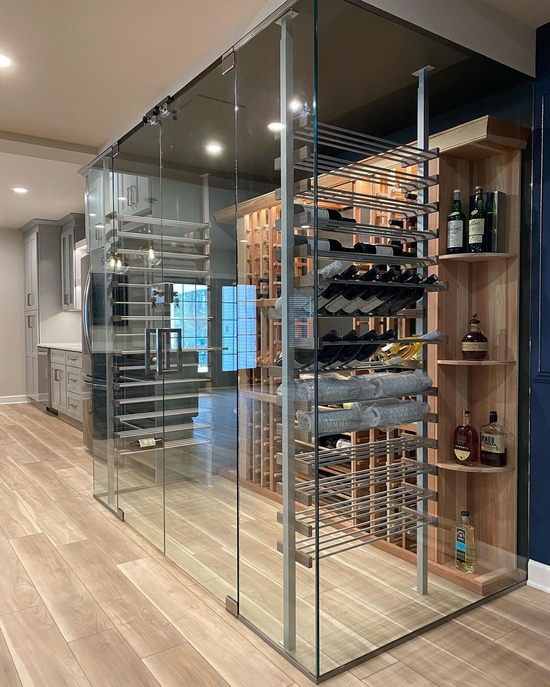 Glass-enclosed wine cellar with various bottle racks, wooden shelves, and bottles displayed. Interior with light wood flooring.