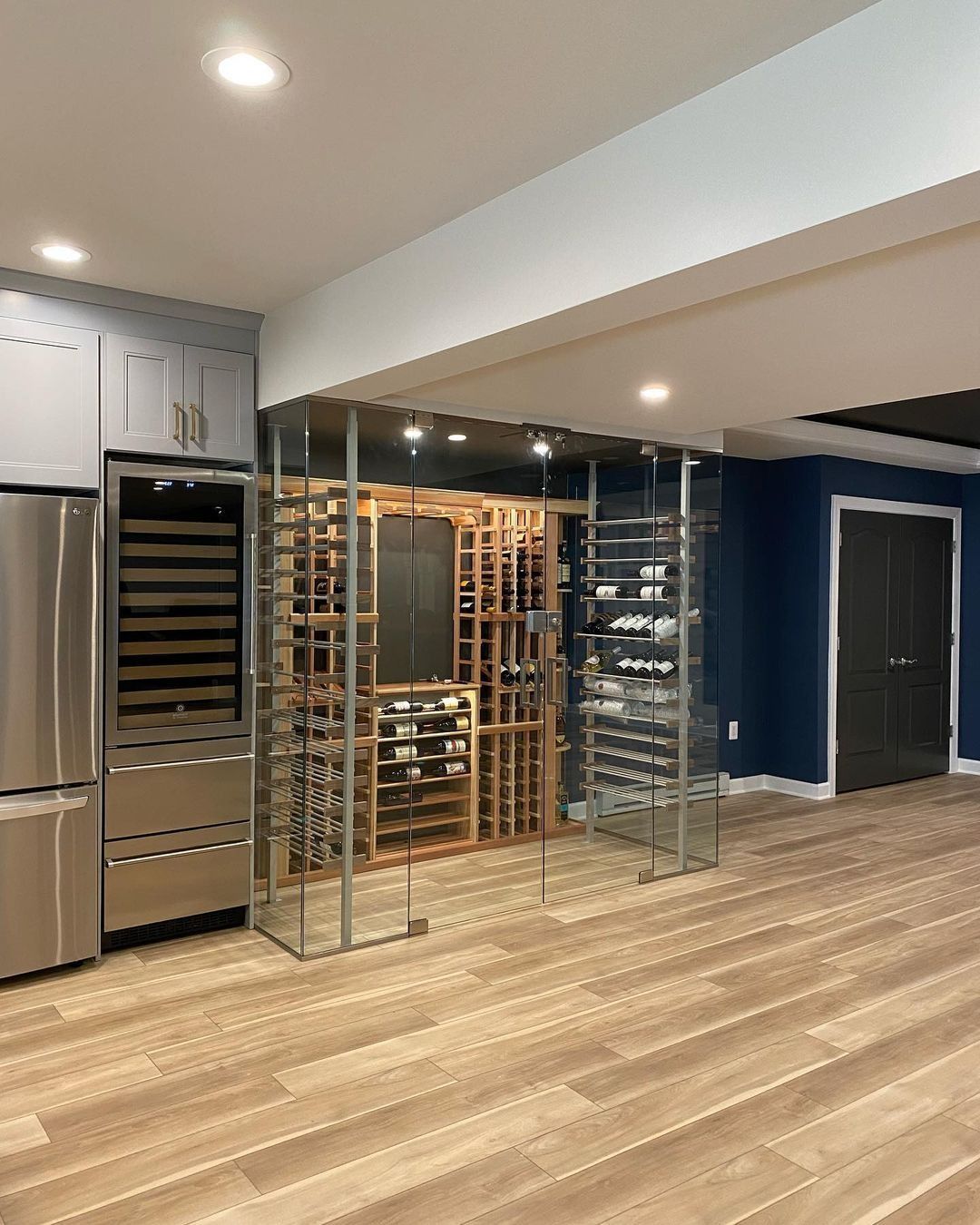 Wine cellar with stainless steel refrigerator, wooden wine racks, and glass shelving in a modern room with wood flooring.