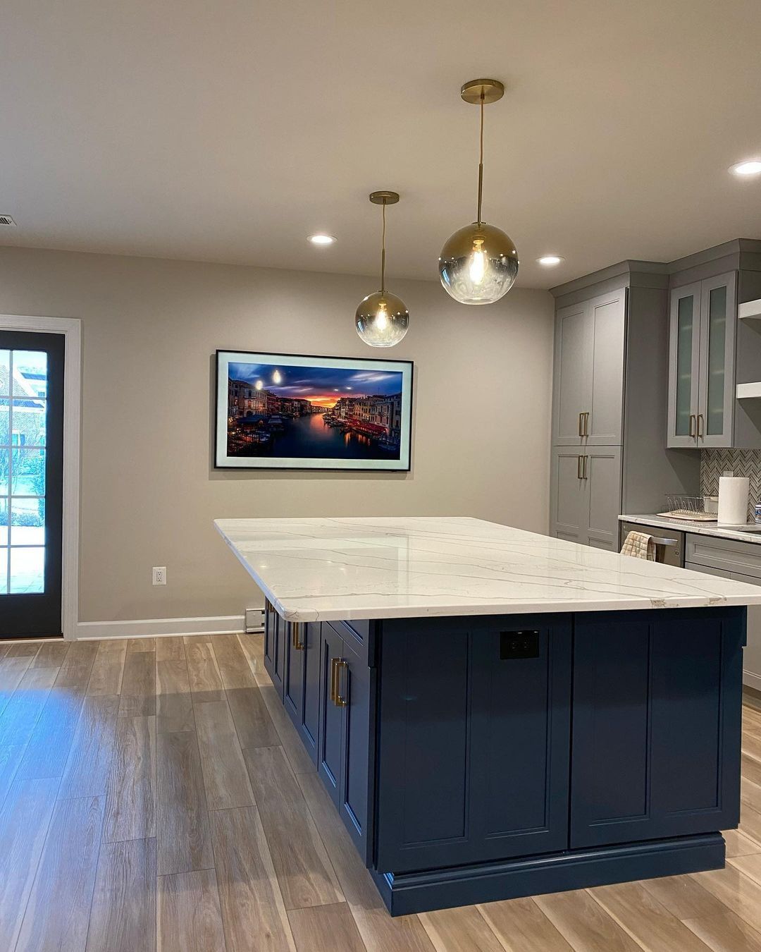 Modern kitchen with blue island, white countertop, gold pendant lights, and TV displaying a city at sunset.