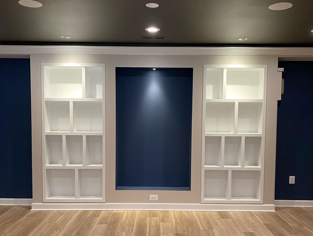 Built-in white shelves flank a dark blue recessed area on a wall; recessed lighting on a dark ceiling.