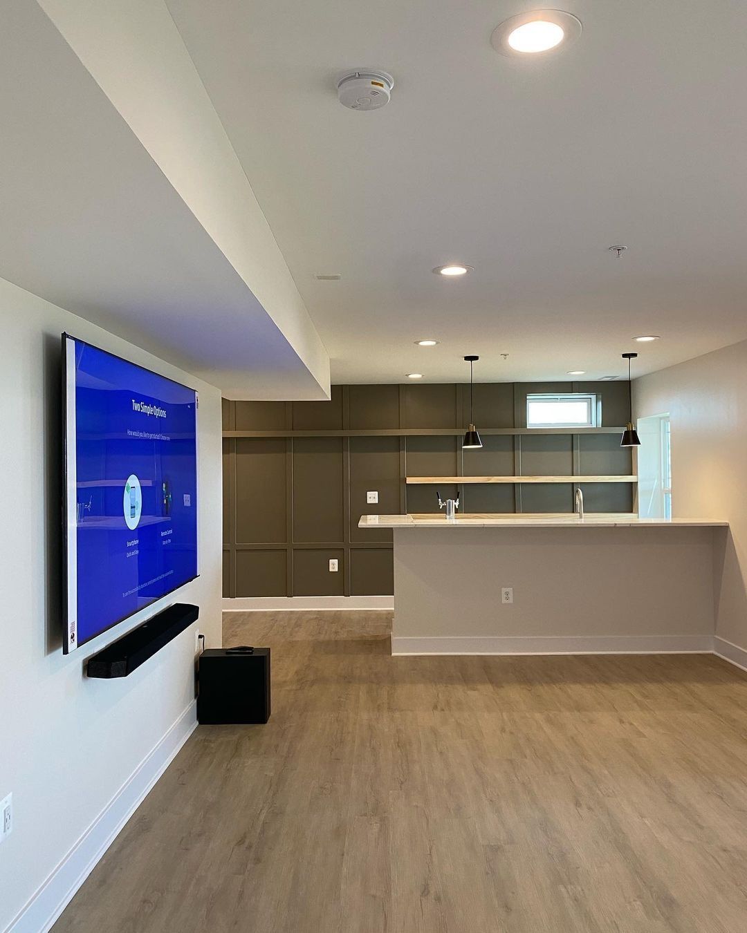 Modern basement with a TV, bar, and light wood flooring; the walls are white, with a darker accent wall.