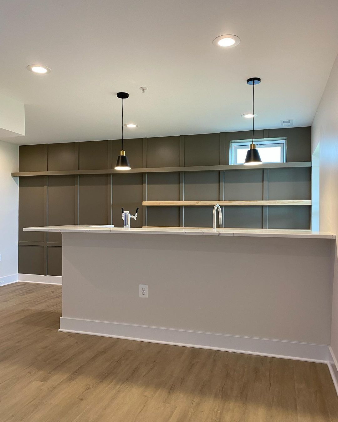 Interior view of a modern kitchen with a gray island, shelves, and dark green accent wall, wooden floors, and pendant lighting.