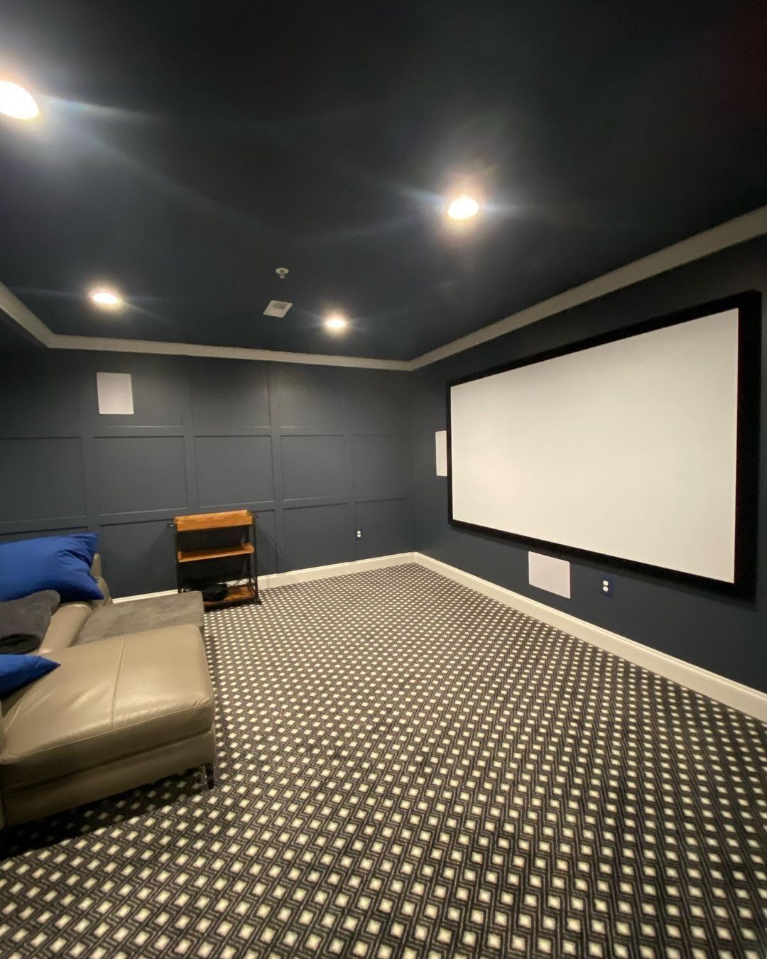 Home theater with navy walls, white screen, and patterned carpet. Beige sofa, wooden shelf, and recessed lighting.