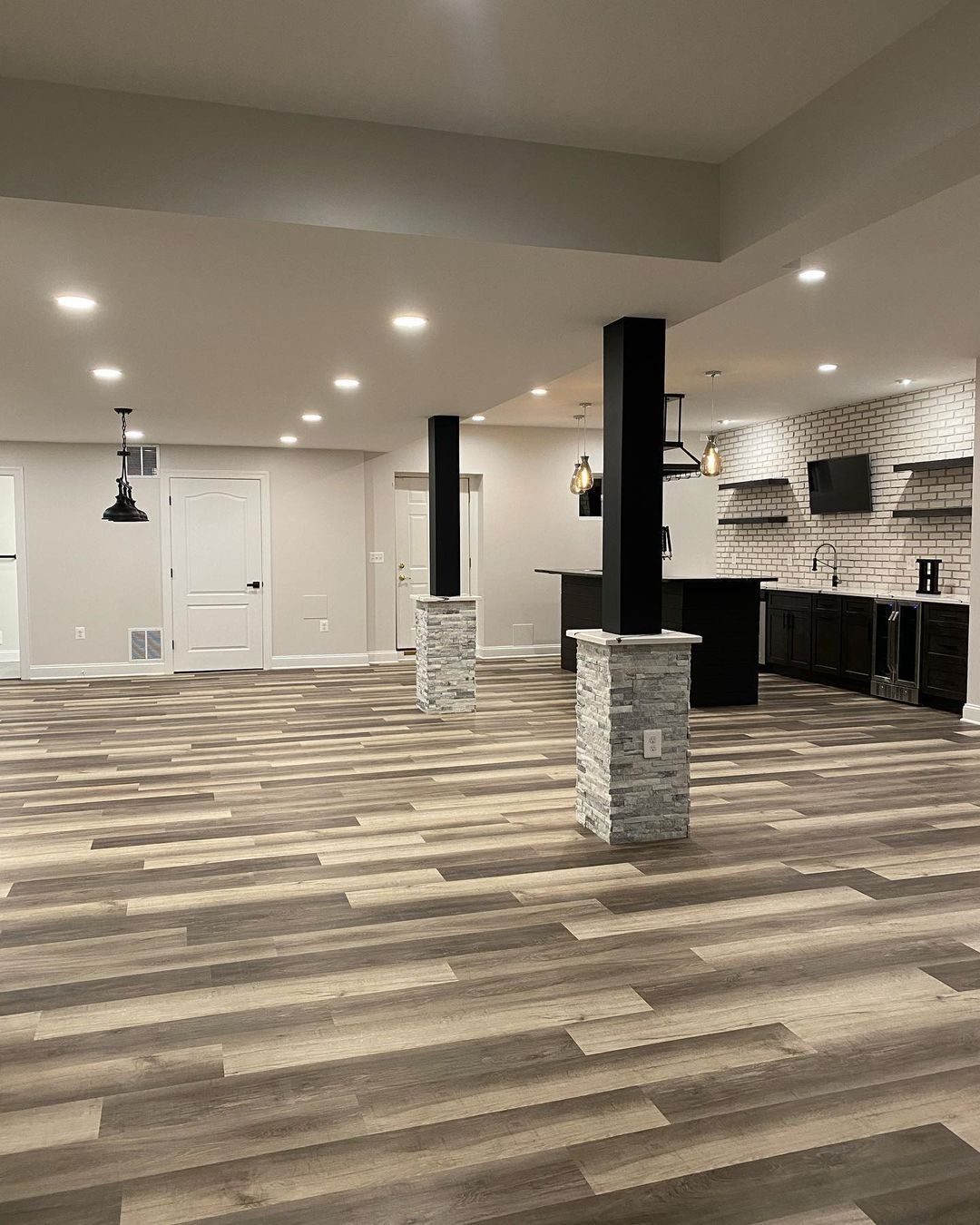 Finished basement with light wood-look flooring, recessed lighting, support columns, and a bar area with brick accent wall.