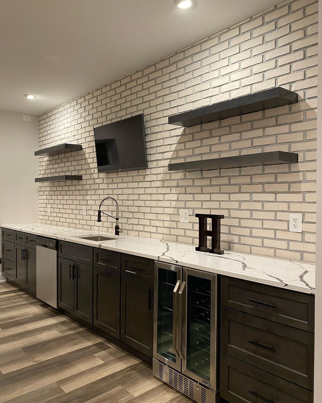 Basement bar with white brick wall, dark cabinets, a countertop, and shelves. A wine cooler and sink are visible.