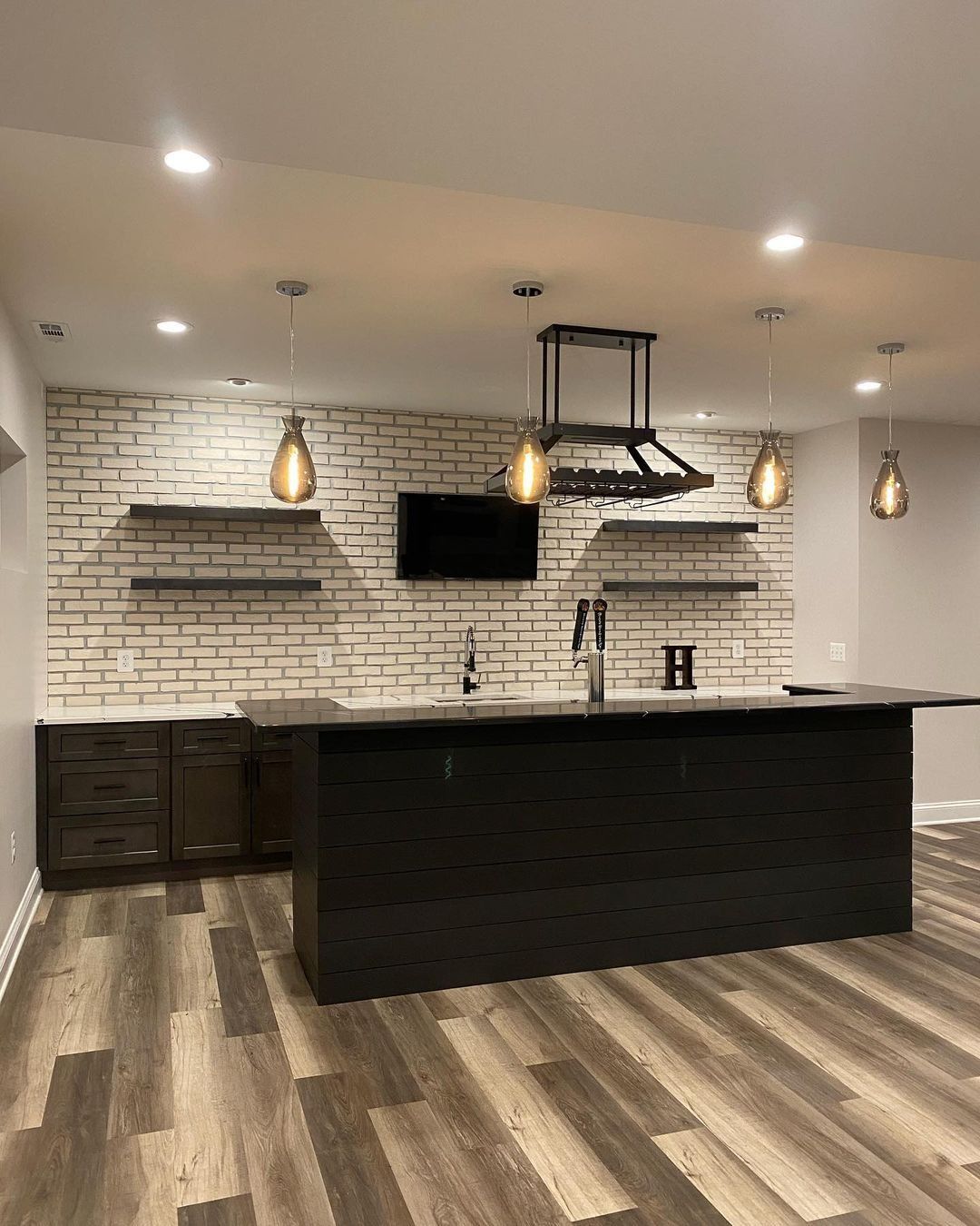 Modern basement bar with brick accent wall, island, dark cabinetry, pendant lights, and wooden flooring.