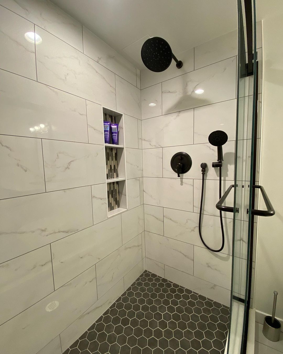 Modern shower with white tiled walls, dark fixtures, and a honeycomb-tiled floor.