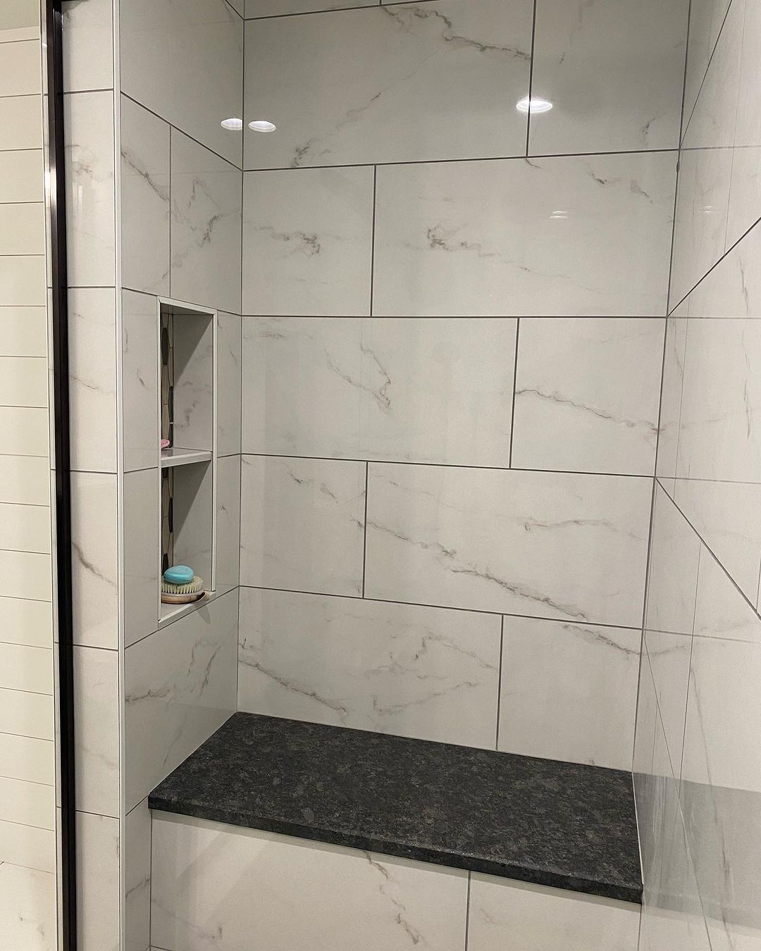 Shower stall with white marble-look tiles, a dark gray bench, and a built-in shelf.