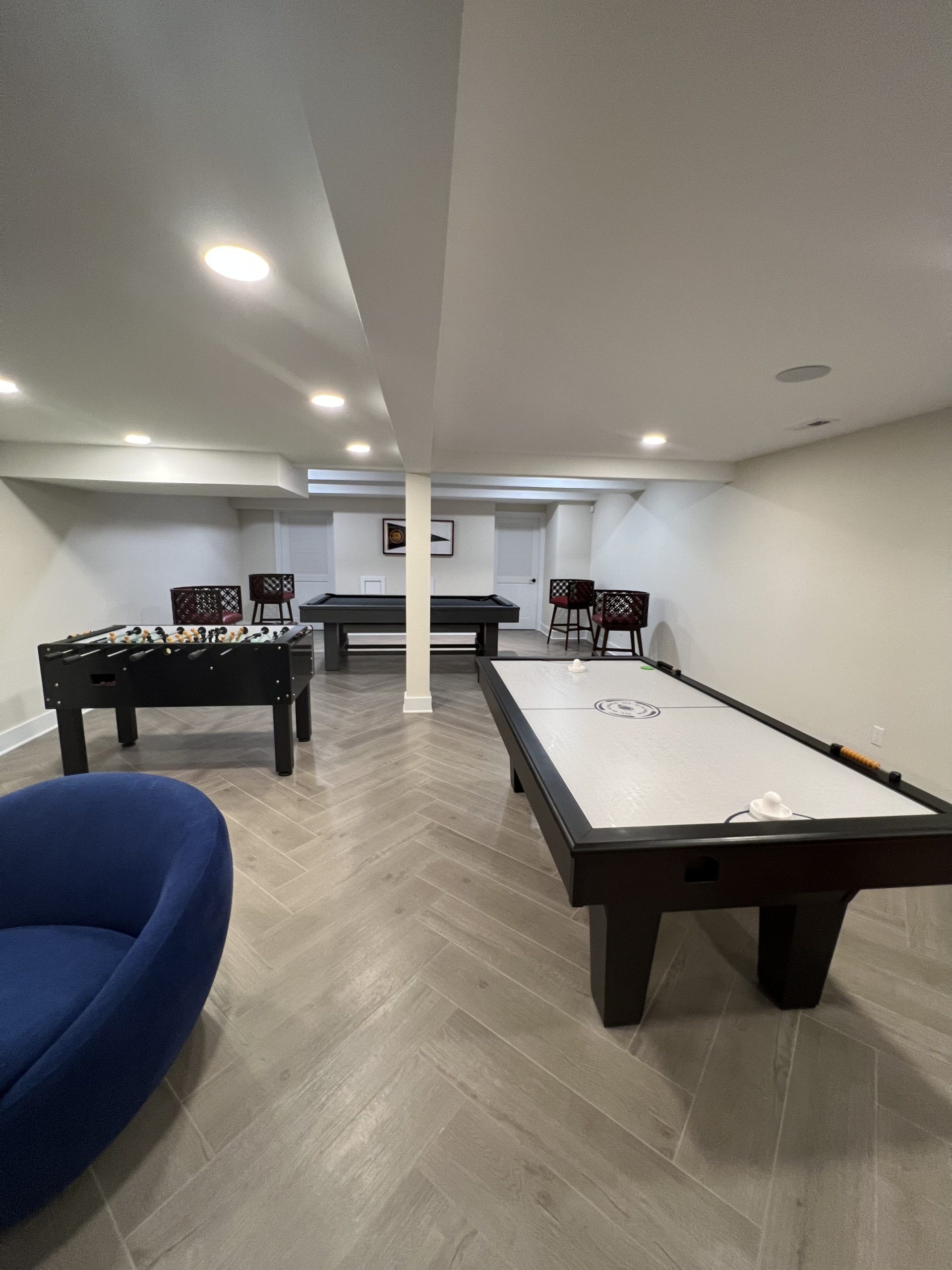 Game room with shuffleboard, foosball, pool table, and blue chair. Herringbone floor, white walls.