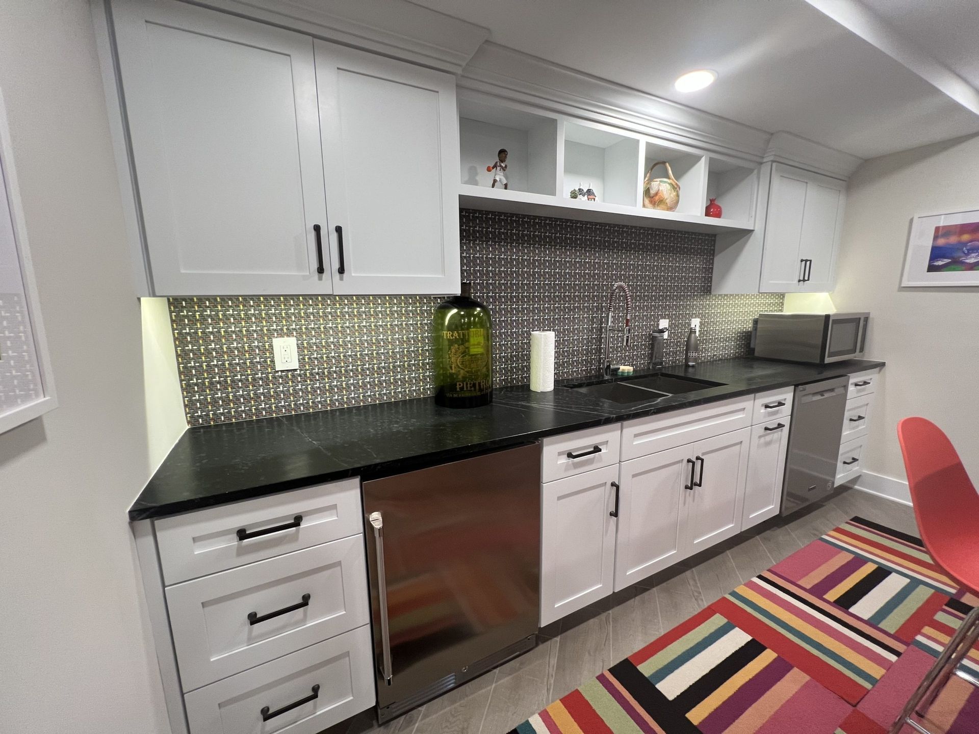 White kitchen cabinets, black countertop, stainless steel appliances, mosaic backsplash, and colorful rug.