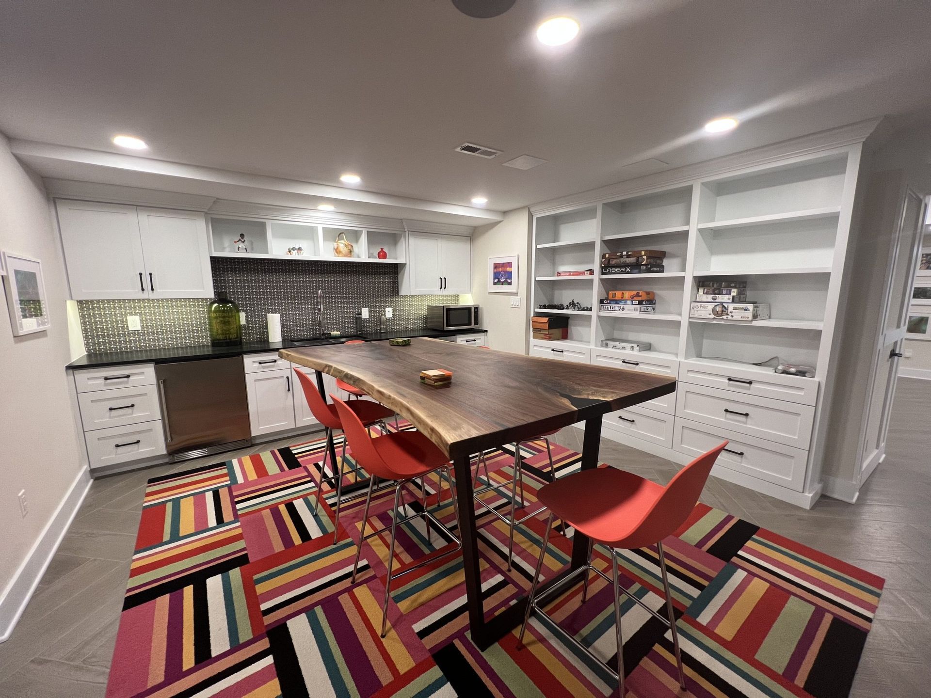 A modern kitchen with a large wooden table, red chairs, and a colorful rug. White cabinets and shelving line the walls.