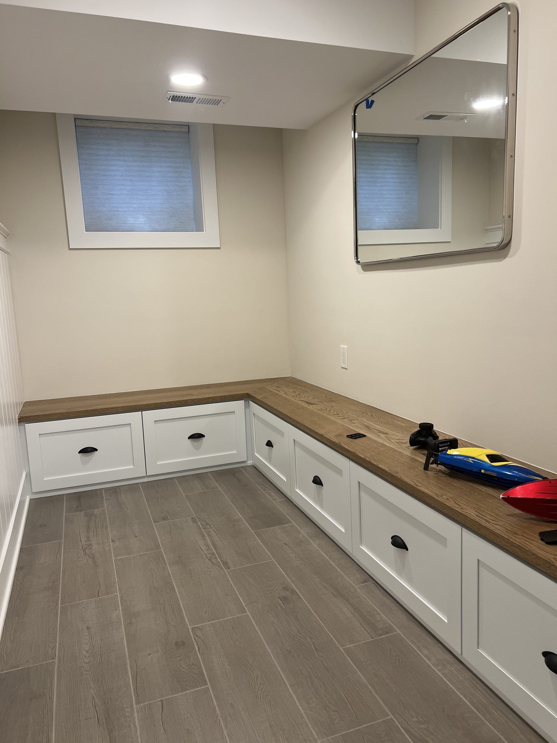 Corner built-in storage bench with wood top, white cabinets, and a mirror on the wall. Window visible.