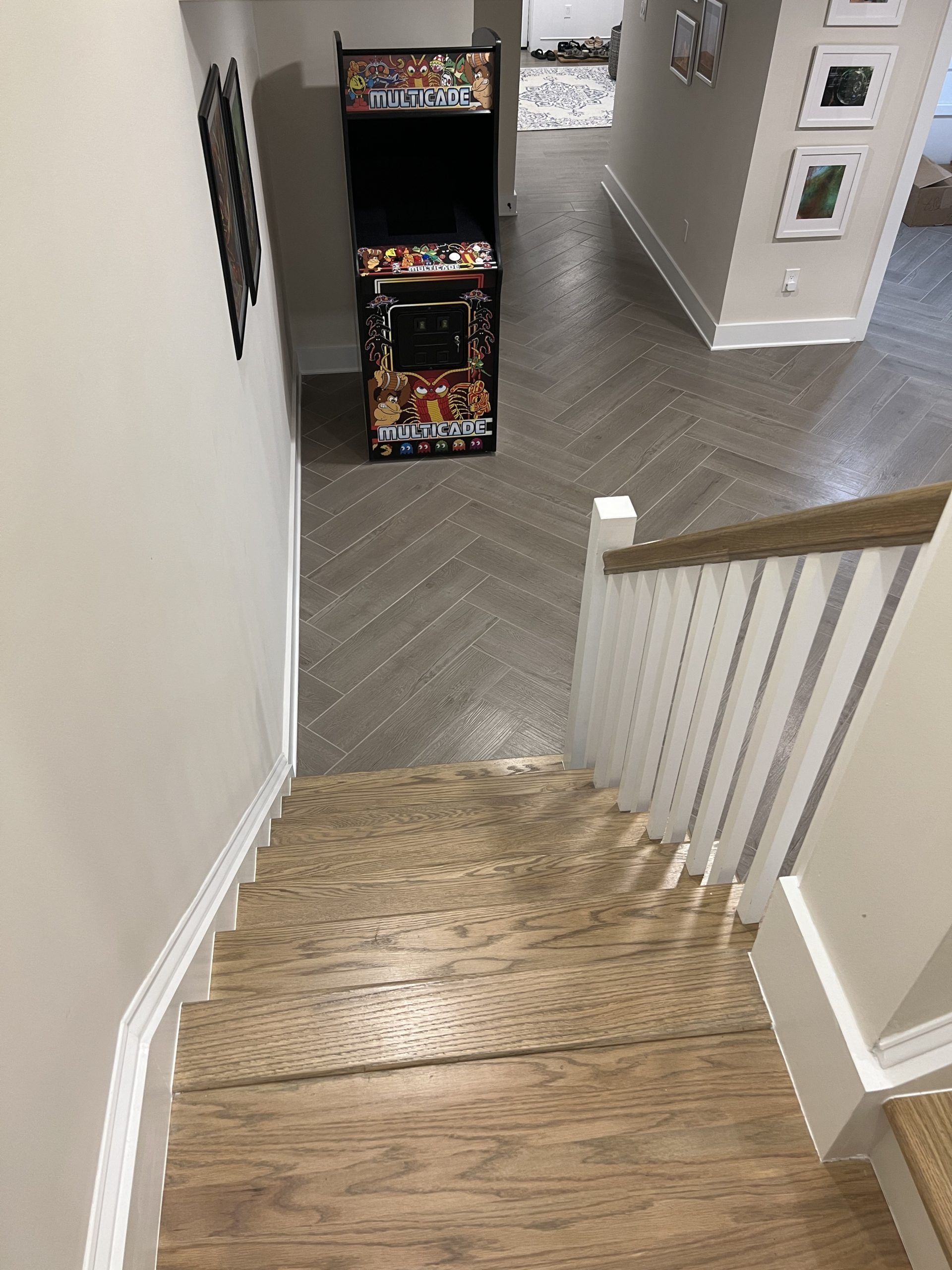 Staircase leading down to a hallway with an arcade game. Herringbone flooring. White walls and railing.
