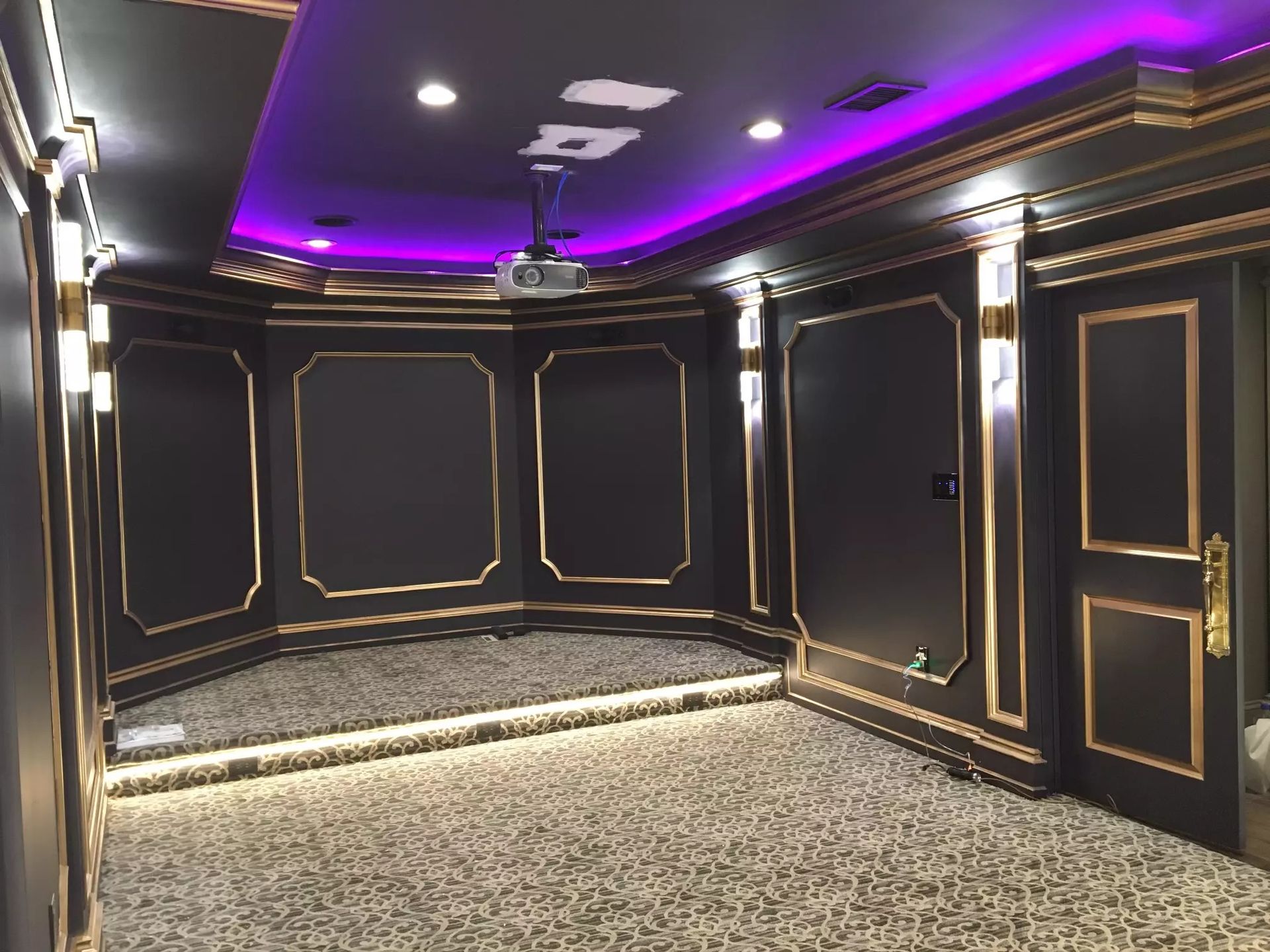 Home theater with black walls, gold trim, purple ceiling lights, and patterned carpet.