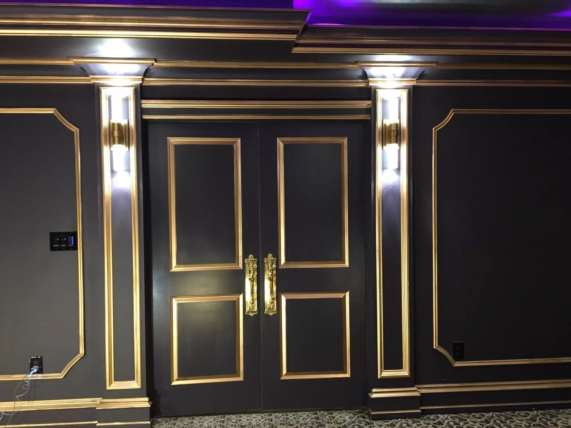 Black double doors with gold trim, flanked by wall sconces, in a dark room with gold crown molding.