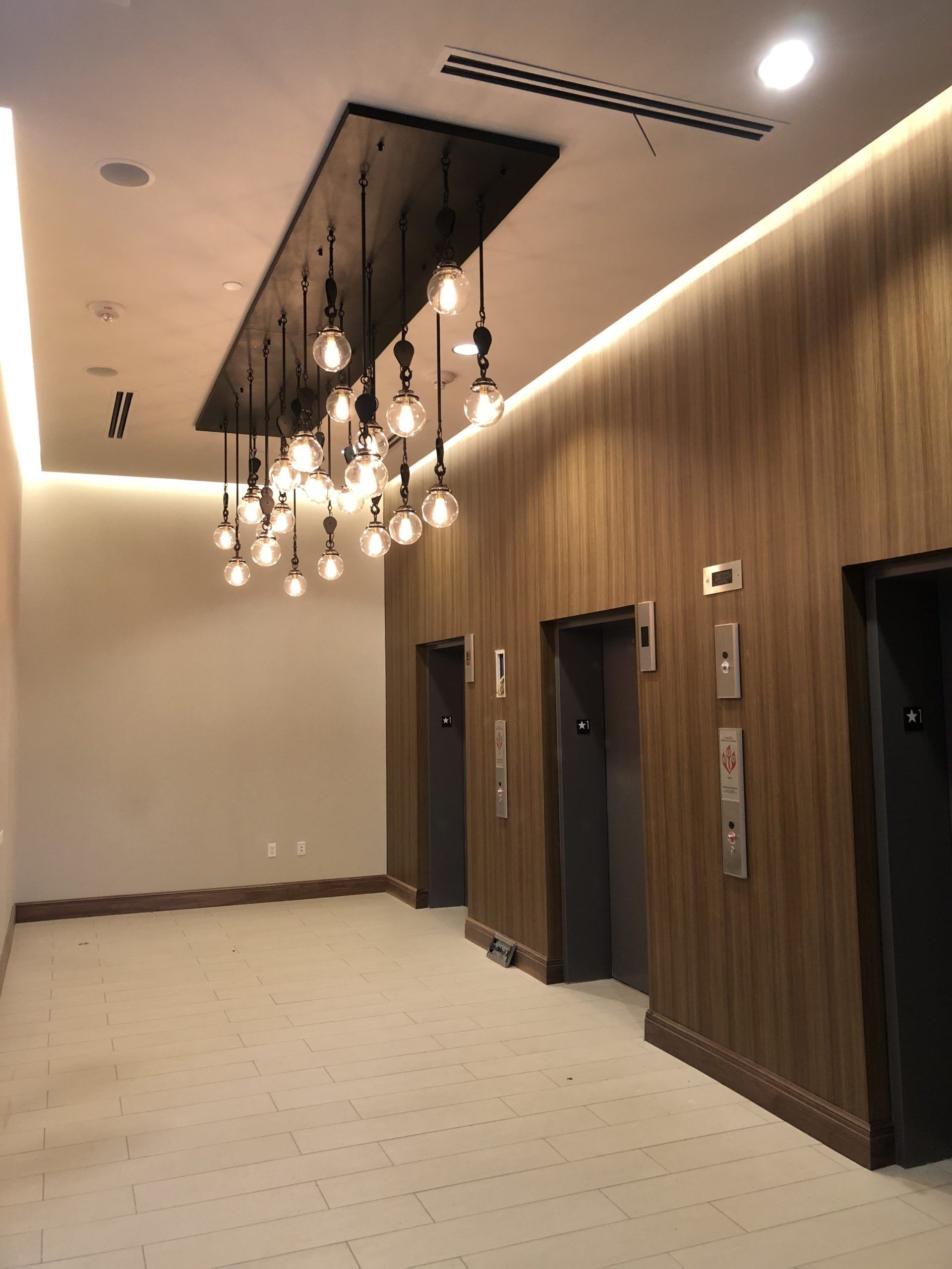 Elevator lobby with wood-paneled walls, three elevator doors, and a chandelier with hanging light bulbs.