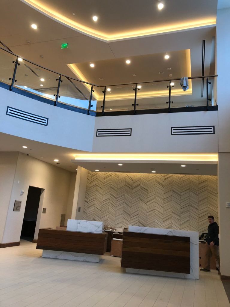 Lobby with reception desks, patterned wall, second-floor balcony, and a person standing behind the counter.
