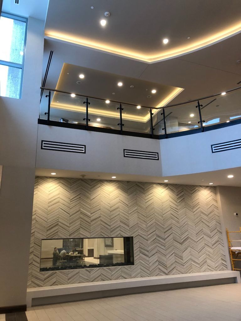 Modern lobby with patterned wall, fireplace, glass railing, and second-floor balcony. White and beige tones.