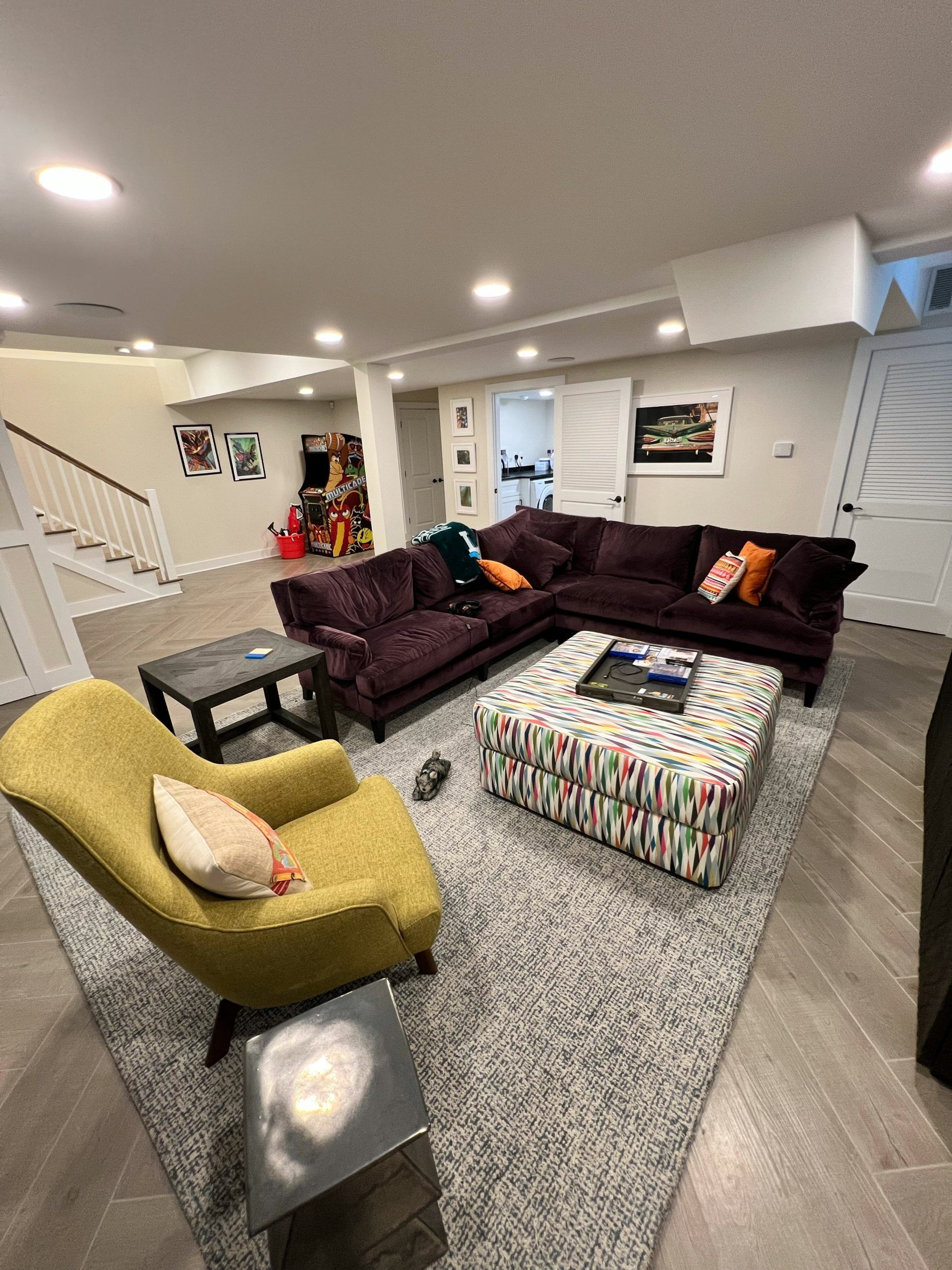 Living room with purple sectional, patterned ottoman, yellow chair, gray rug.