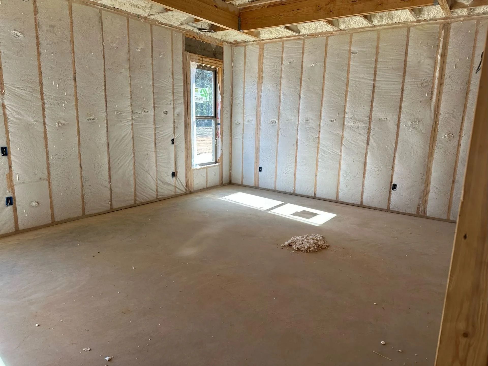 Unfinished room with insulation between wooden studs, window, and concrete floor.