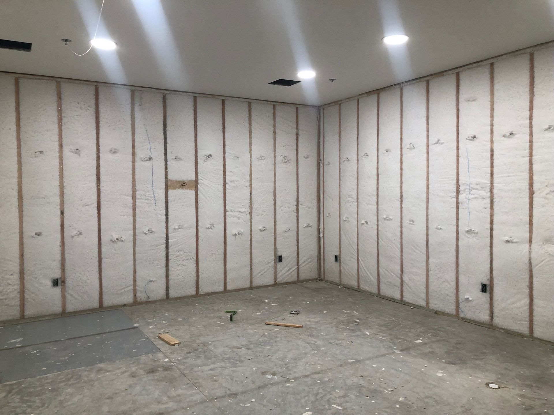 Interior room with insulation sprayed between wooden studs on walls. Concrete floor and recessed lights.