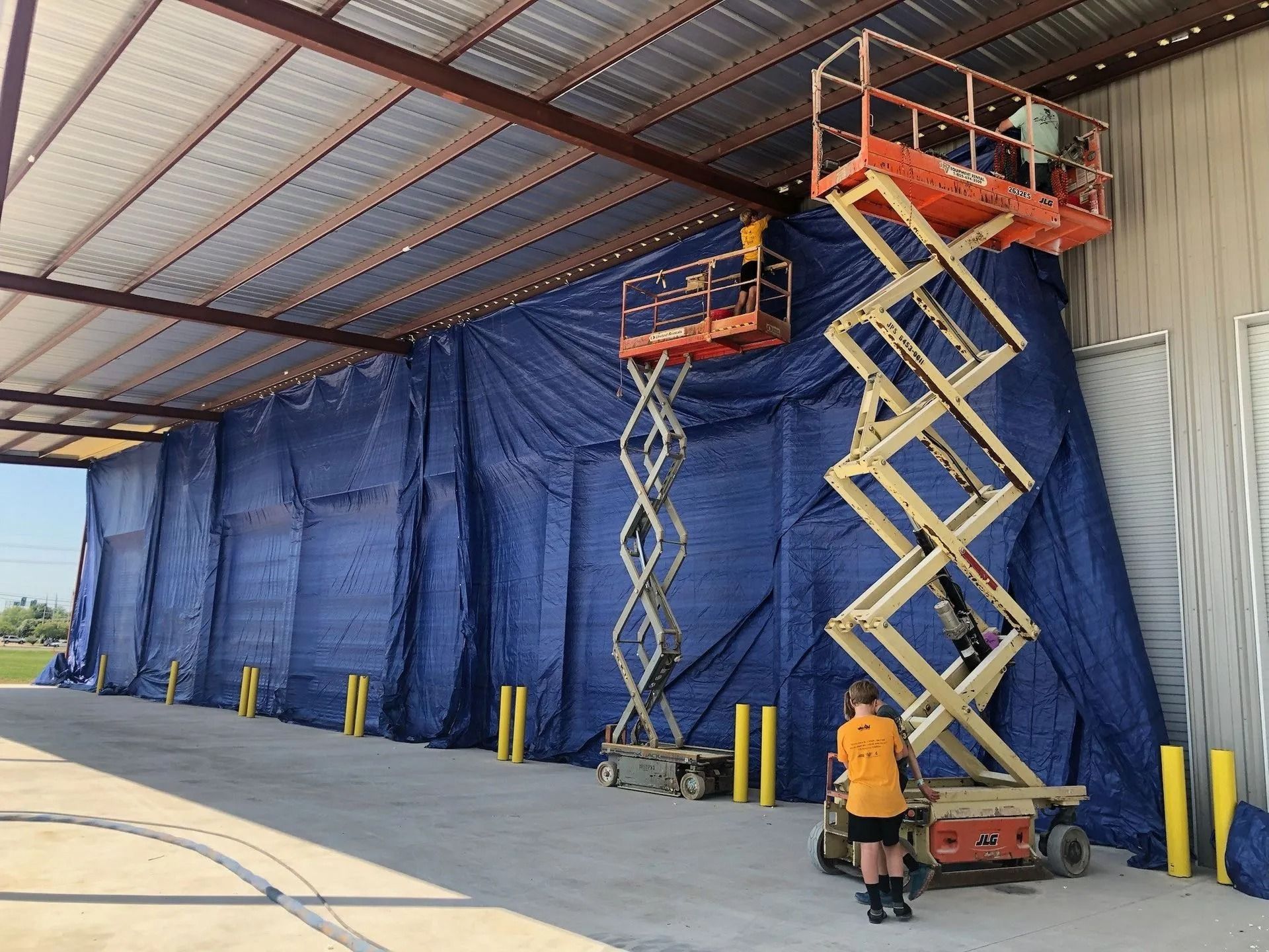 Workers on scissor lifts attaching blue tarps to the side of a building under a metal awning.