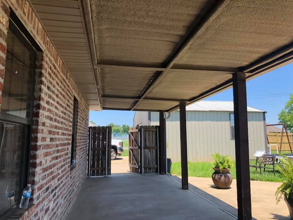 Covered patio with tan shade panels, brick wall, and concrete flooring. A gate leads to a yard with buildings.