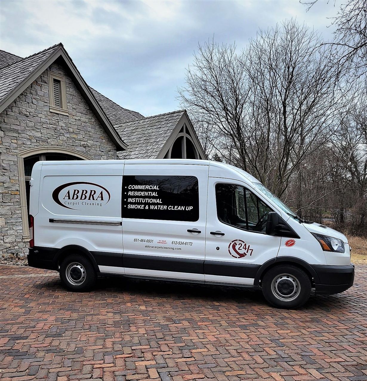 White service van parked on a brick driveway in front of a stone house and leafless trees