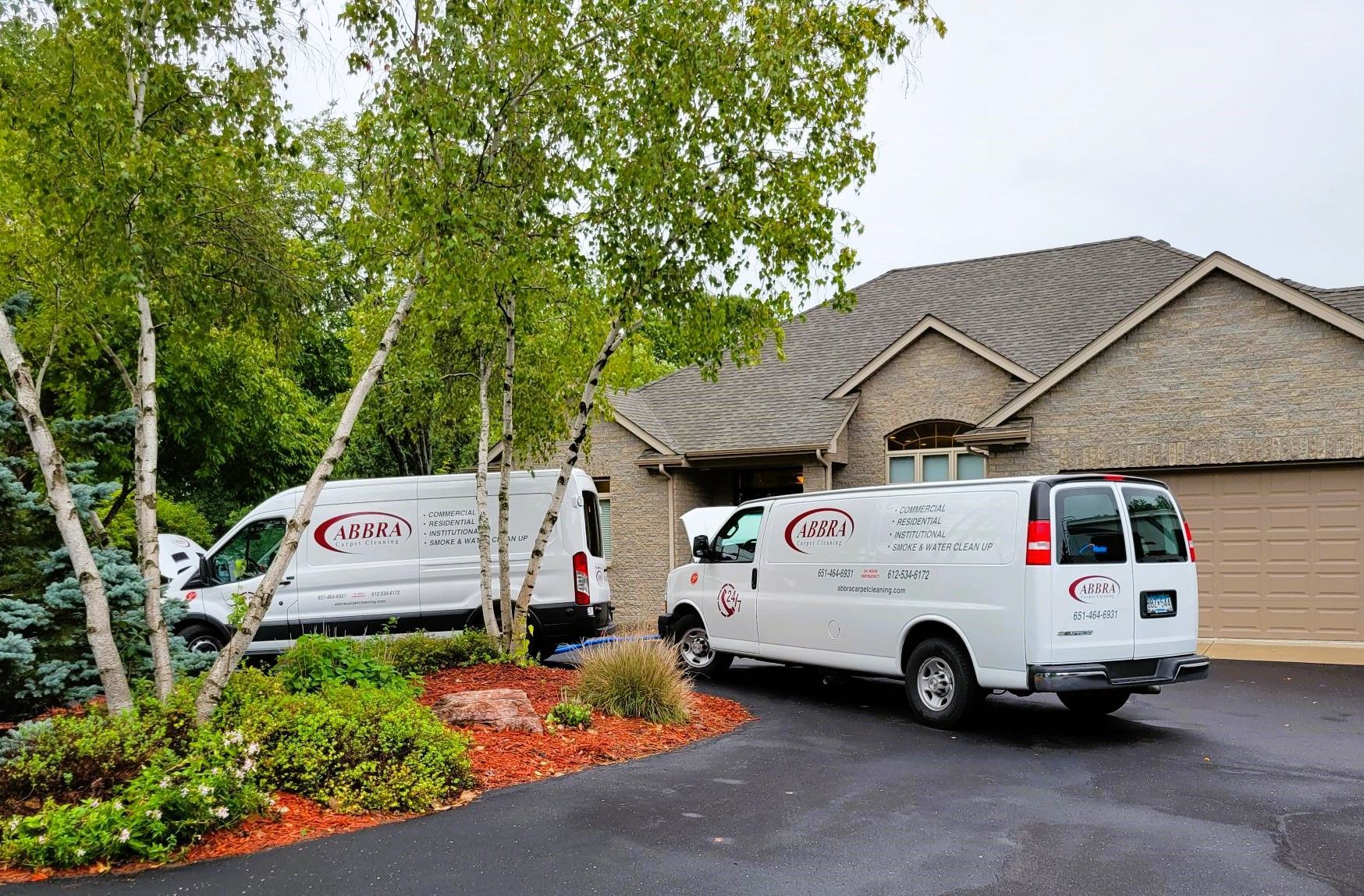 White service vans parked in front of a brick house and wooded landscaping on a wet driveway