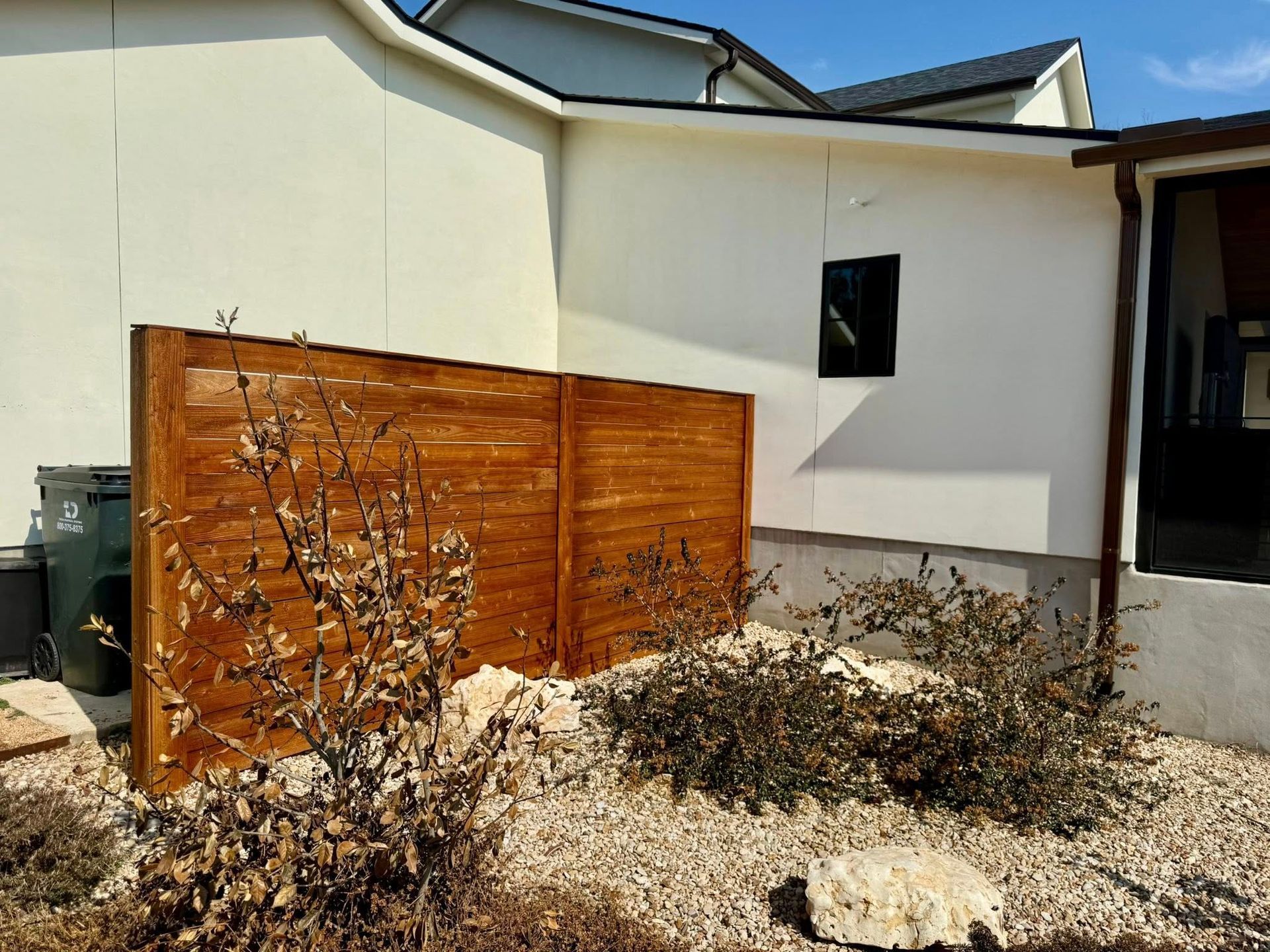 Wooden privacy fence against a white building; gravel landscaping with shrubs and a rock.
