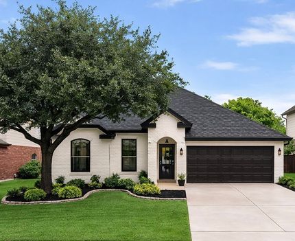 Suburban house with cream brick exterior, dark garage, manicured lawn, and large front tree under blue sky