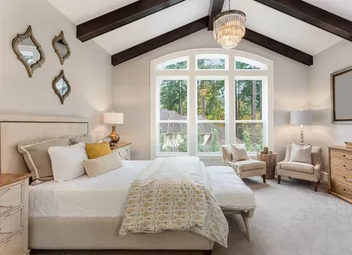 Bedroom with large arched window, white walls, and dark wooden beams. Bed with white linens, chairs, and ornate mirrors.