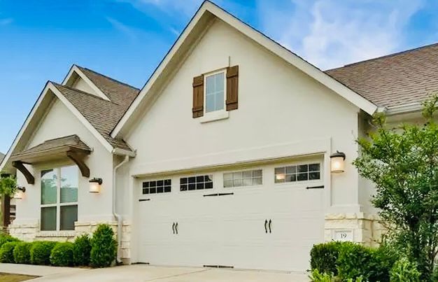 White stucco house with brown roof, shutters, and garage door; blue sky.