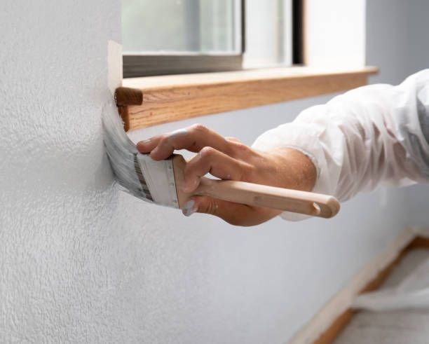 Person painting a white wall with a brush near a wooden window frame.