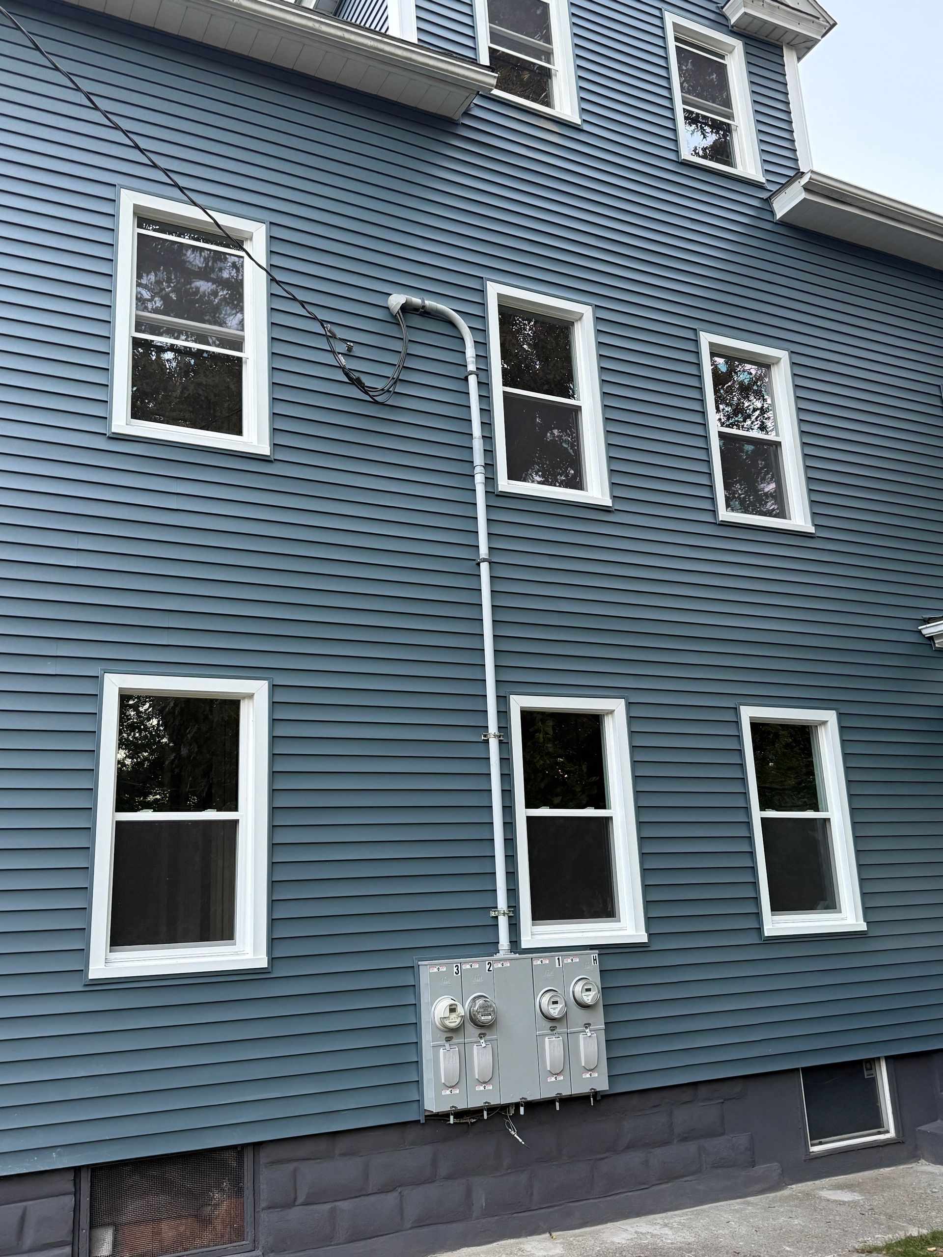 Blue-sided building with white-framed windows, electrical boxes, and a conduit running down the side.