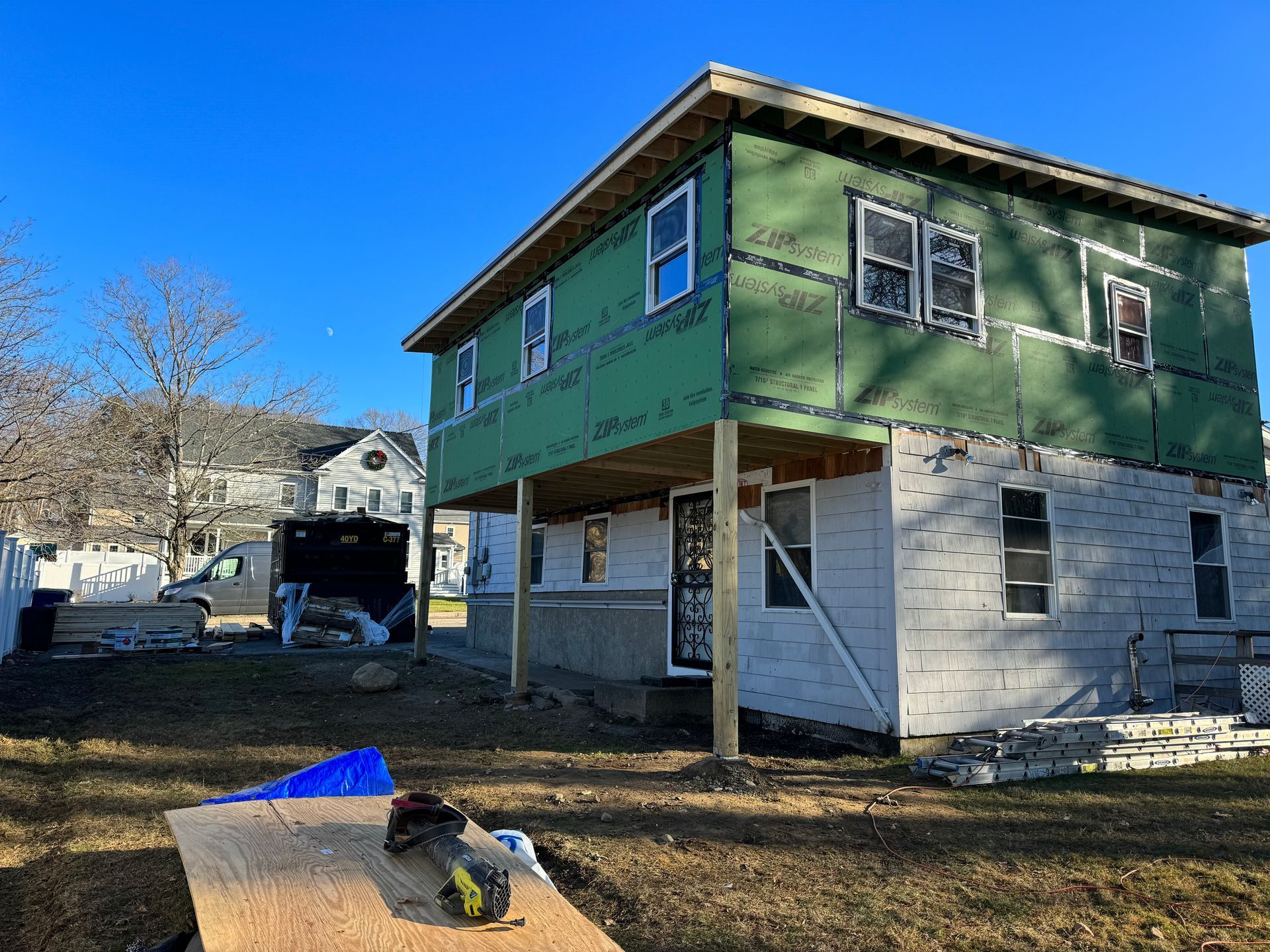 Two-story house under construction, green siding, wood beams supporting the upper level, sunny day.