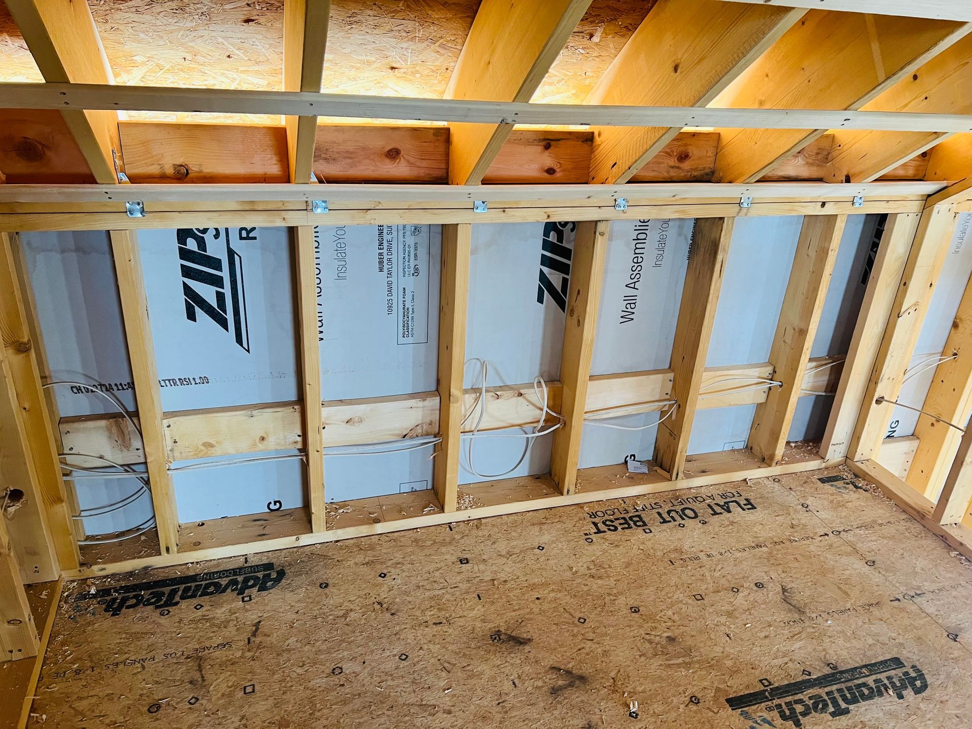 Interior view of a room under construction with exposed wooden framing and grey wall sheathing. Electrical wires visible.
