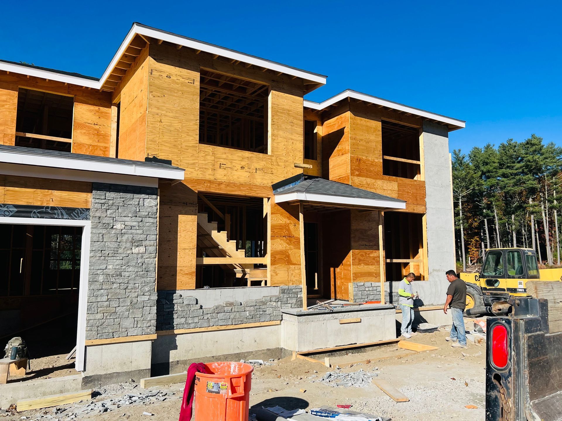 Construction of a two-story house with wood framing and stone accents; two workers on site, clear sky.