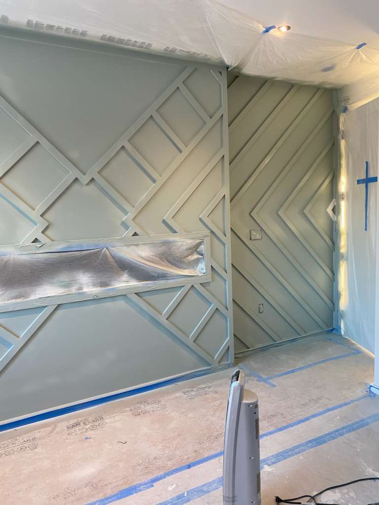 Room with light green geometric wall paneling, blue painter's tape, and a fan.